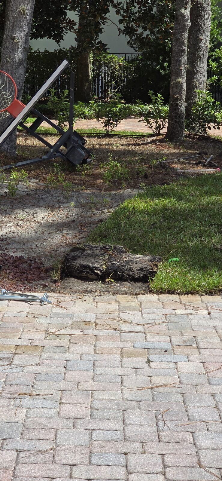 Old portable basketball hoop removed and laid on its side beside a freshly dug-out hole on a paver driveway