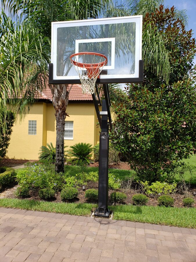 In-ground basketball hoop set at the edge of a paver driveway in front of a yellow Florida home