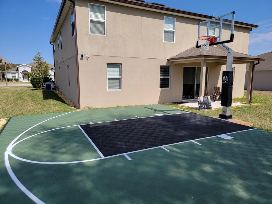 Green and black sport-court tiles with white lines and an in-ground hoop behind a two-story home