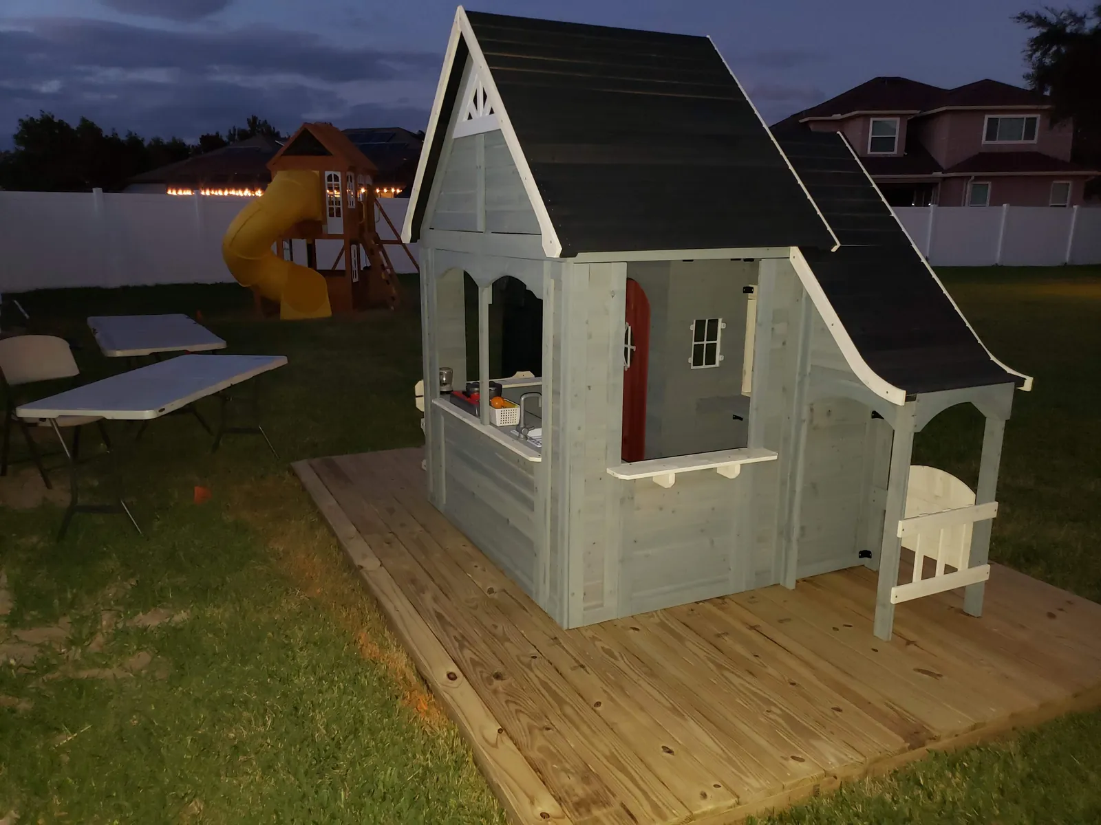 Wooden backyard playhouse with shingled roof and porch finished at dusk on a pressure treated deck in Florida
