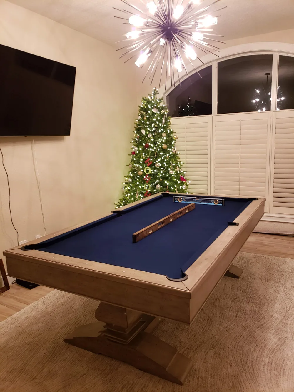 Two machinist levels laid across the navy blue felt of a rustic pool table during slate leveling in a Florida game room, decorated Christmas tree glowing behind it
